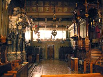 Photograph of room with ornate columns and pews along both sides, a raised platform on the right side, and several chandeliers. 