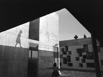 Photograph of exterior of modern apartment building taken from below an overhang showing a young woman walking toward a wall in front of her, and shadow of another woman reflected on the side of the building on the left.