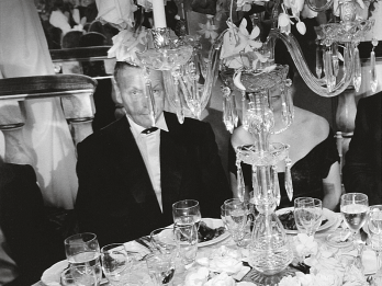 Photograph featuring man seated at a dinner table covered with dishes with his face partially covered by a chandelier.