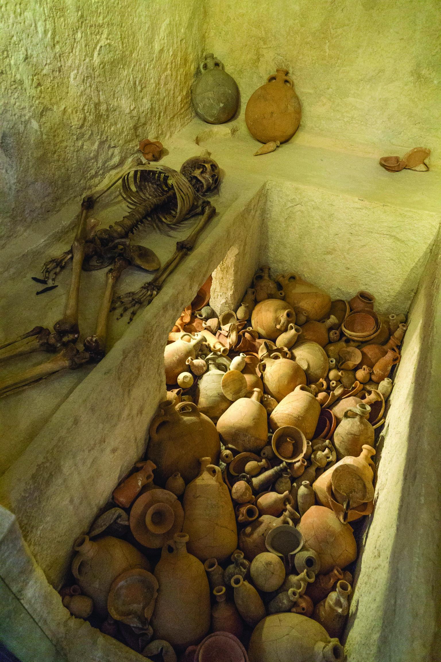 Interior room with ledges, one of which holds a skeleton, and a floor covered with pottery.