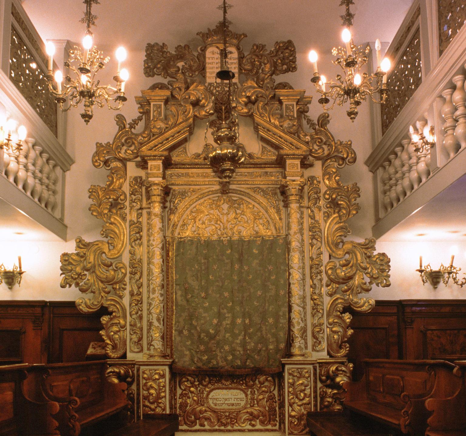Photograph of room with large carved Torah ark with pews on either side of room and screened second-floor balcony. 