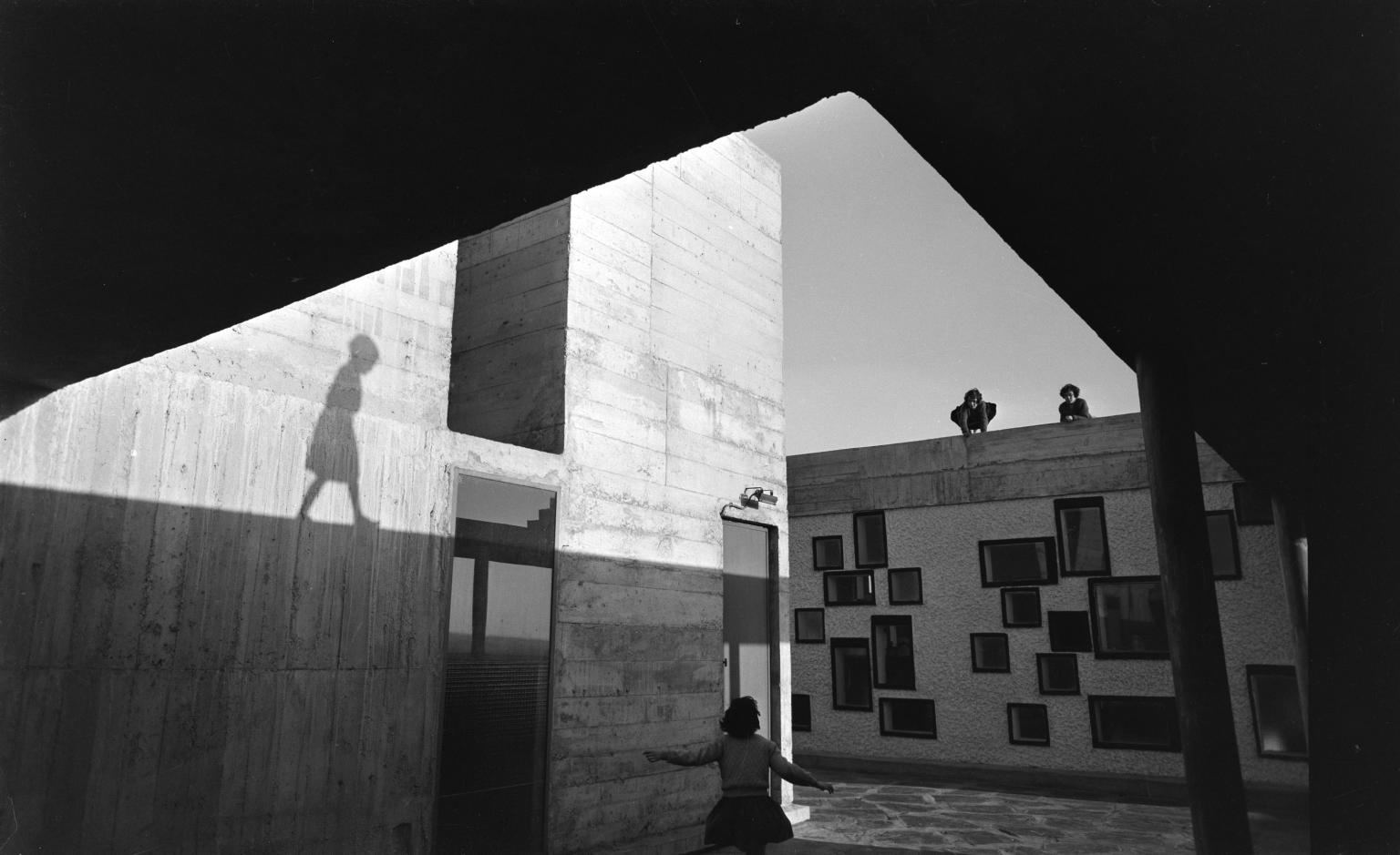 Photograph of exterior of modern apartment building taken from below an overhang showing a young woman walking toward a wall in front of her, and shadow of another woman reflected on the side of the building on the left.