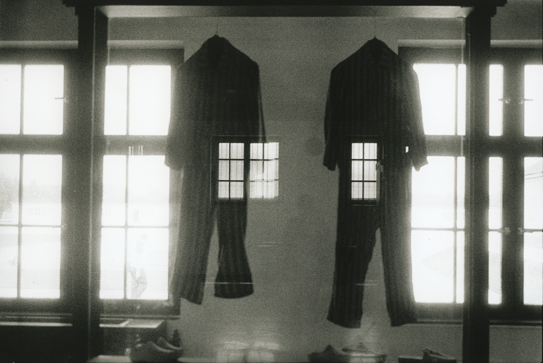 Photograph depicting two jumpsuits hanging between two large windows superimposed over an image of a human figure standing with their back turned. 