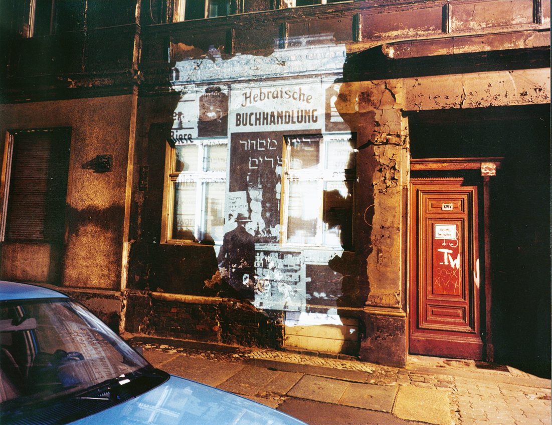 Photograph of blank city wall with projection of storefront over it and car in foreground.