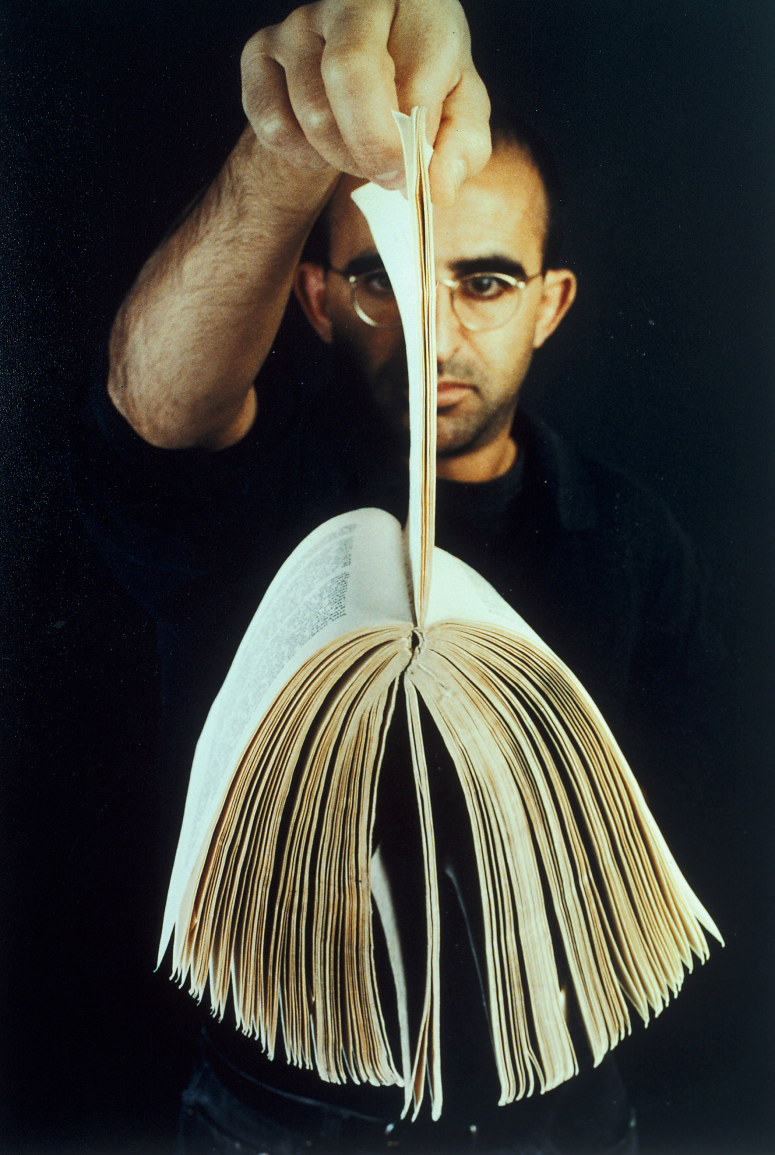 Photograph showing man facing camera and holding up a book by its pages.