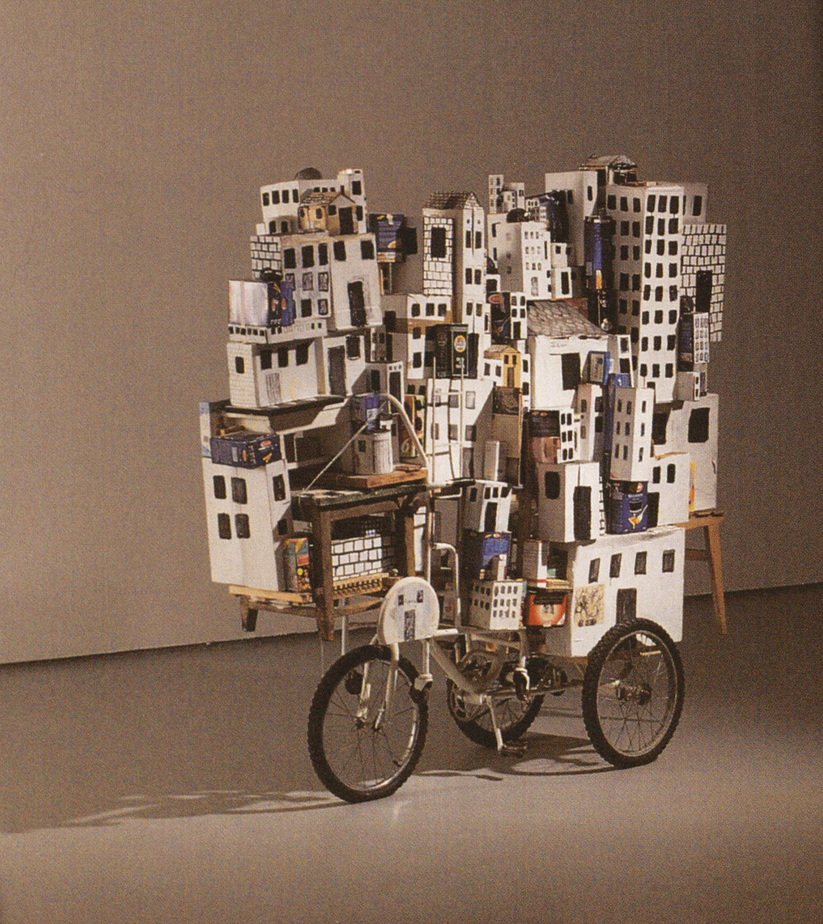 Mixed media work featuring stack of buildings on bicycle. 