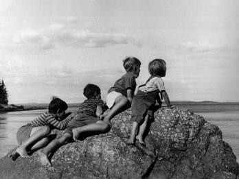 Photograph of four boys perched on a rock overlooking water and facing away from viewer.