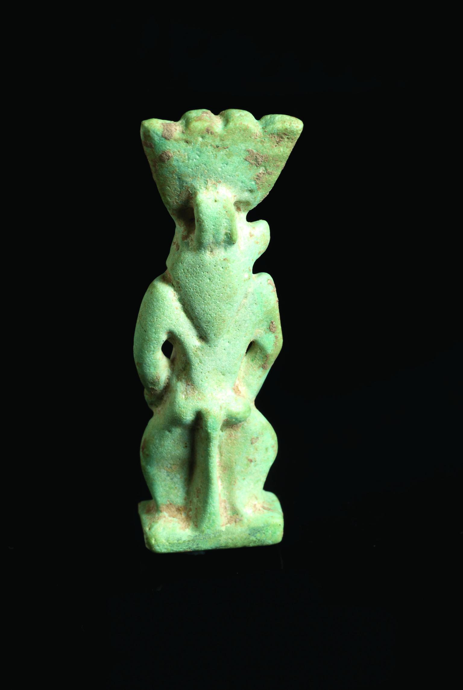 Frontal and back faience amulet of figure who is nude with feathered headdress and a grotesque face and beard.