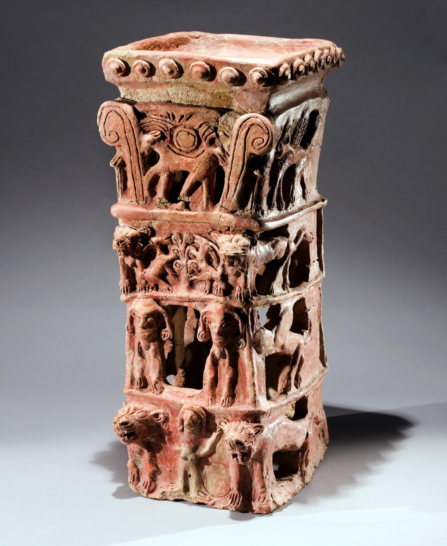 Ceramic offering stand with flat top and four layers of decoration: bottom layer of nude female with raised arms flanked by lions, second layer of two winged figures, third layer of goats eating leaves off tree, and fourth layer of four-legged animal with winged sun disk and palm fronds. 