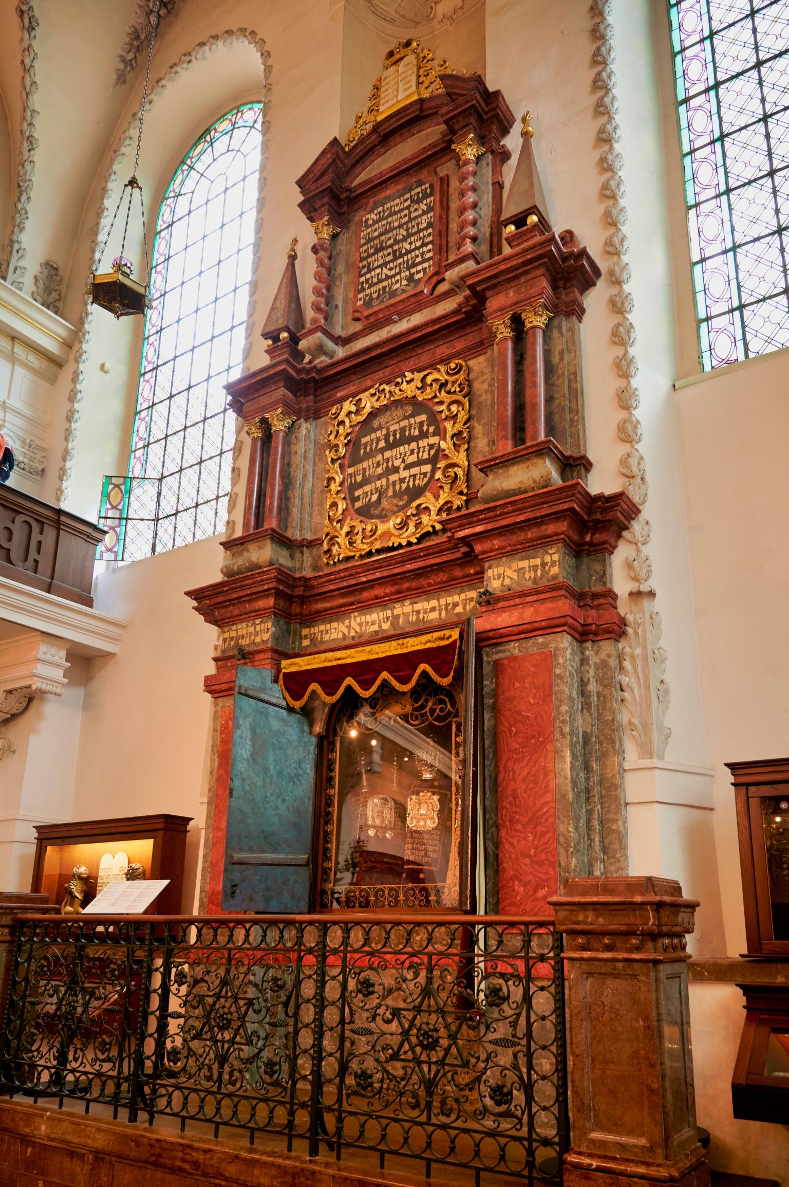 Photograph of tall ark holding two Torahs built into interior wall with Hebrew writing and surrounded by short fence with Stars of David. 