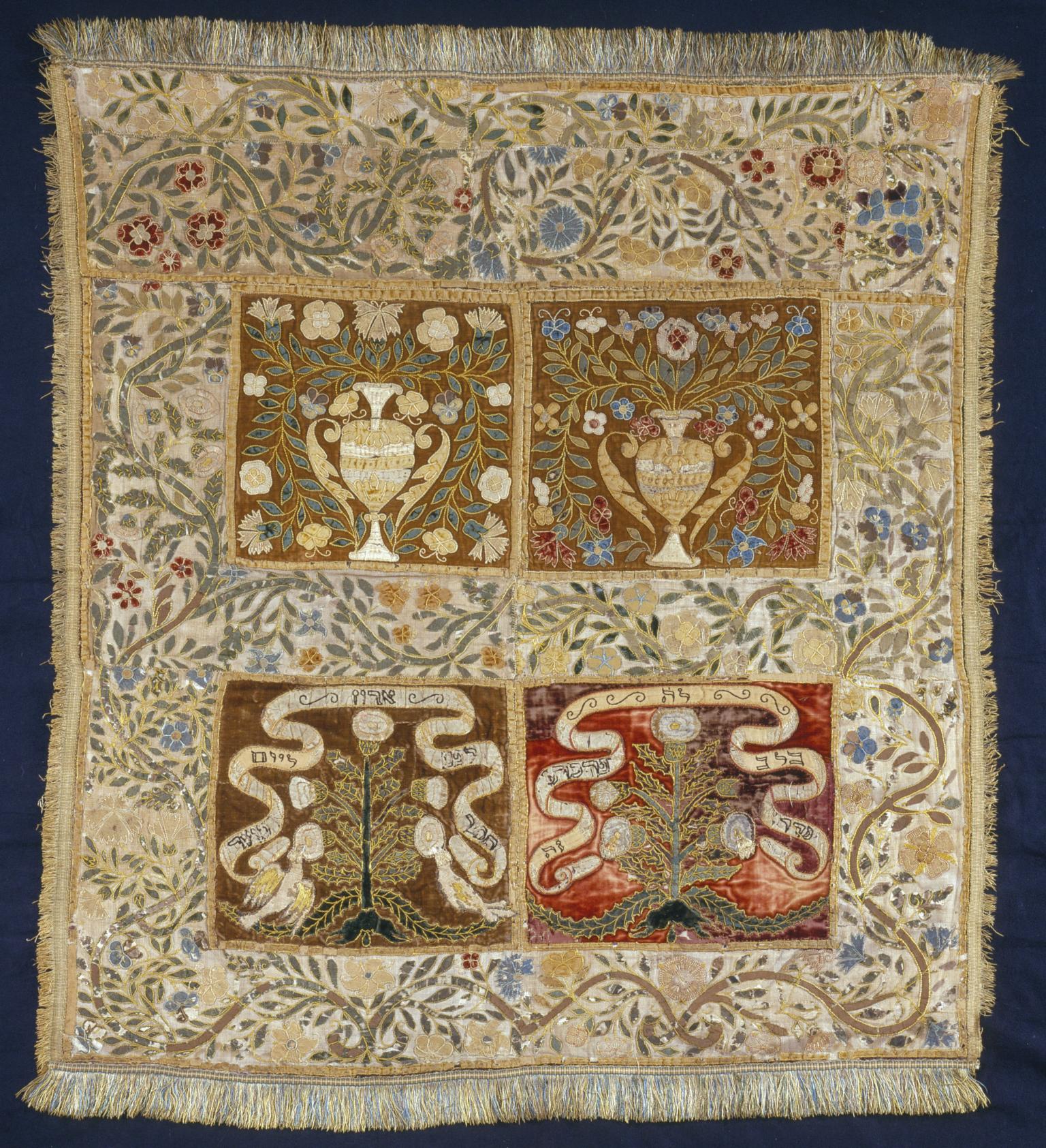 Cloth embroidered with floral border, and four central panels, the top two with pitchers, vines, and flowers, and the bottom two with trees and ribbons with Hebrew text. 