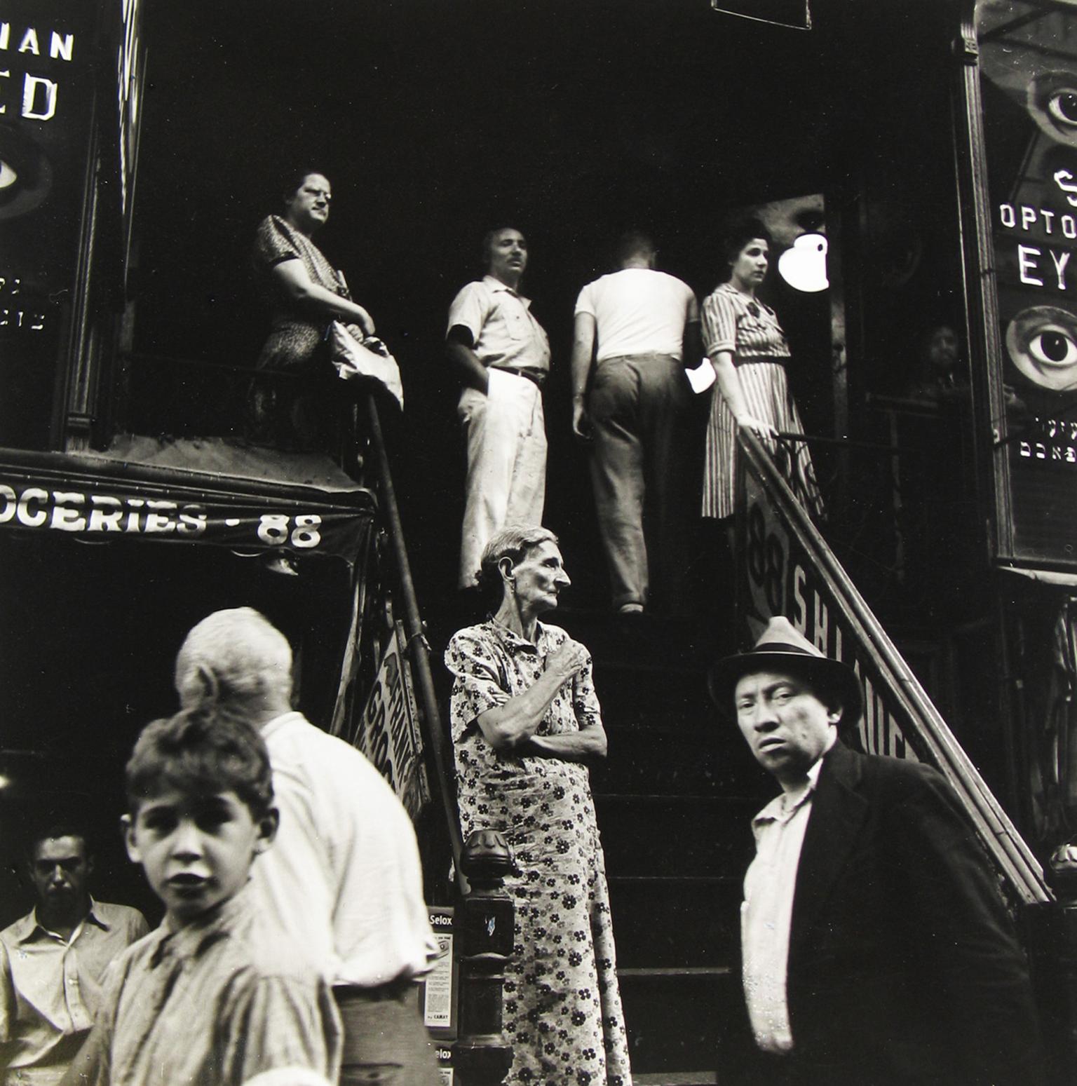 Photograph of people standing outside along stairwell and storefronts.