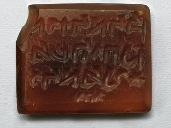 Photo of a rectangular red stone block with a beveled edge and chipped corner, inscribed with reversed Hebrew writing in three lines followed by a small word.