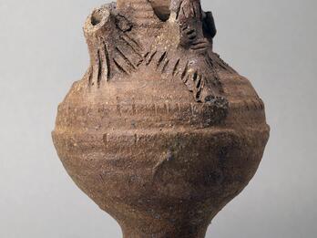Earthenware vessel with a round body and flared base, with sculpted elements, some broken off, at the top, including a humanoid figure.
