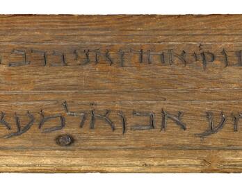 Rectangular mounted wooden plaque with two lines of carved Hebrew script.