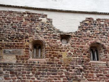 Photo of a weathered stone wall with two arched windows, a small circular window, several irrecular patches, and white-plastered brick along the top.