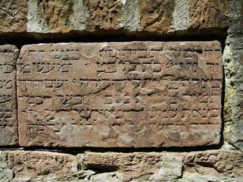 Photo of a stone plaque with weathered, engraved Hebrew text, mounted into a brick wall.