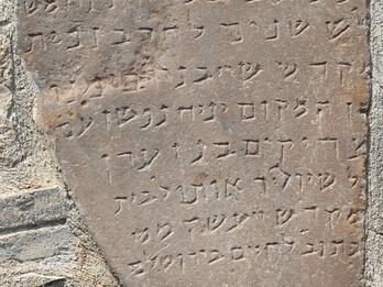 Photo of a weathered, irregular stone tablet, mounted into a brick wall, with a Hebrew inscription conforming to its shape.