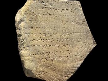Hexagonal, weathered stone tablet inscribed with Hebrew text and two simple ritual candelabrums.