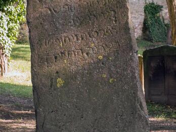 Photo of a severly weathered rectangular gravestone standing upright in a cemetery, with five lines of delineated Hebrew inscription on the top half.
