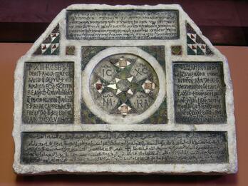 Photo of a hexagonal stone slab engraved with thickly-borded inscriptions in Latin, Hebrew, and Arabic, among which are geometric decorations, including, centrally, a circular pattern with four Latin letter pairs.