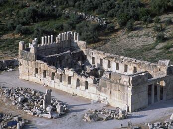 Aerial view of the ruins of an ancient stone building with columns and walls partially standing, surrounded by scattered stones and greenery in the background.