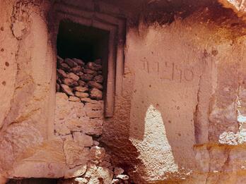A stone doorway carved into a rock wall, filled with loose stones. There is an inscription to the right of the doorway. The rock surface is rough and bathed in sunlight.