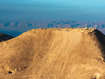 Aerial view of mountain with ruins on top, surrounded by desert landscape, and a sea in the background.