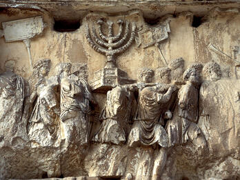 Ancient, weathered stone relief showing people in robes carrying a large menorah and other objects in a procession.