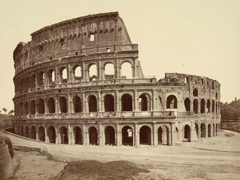 Black-and-white photo of a large amphitheater, showing its ancient stone arches and partial ruin. The large amphitheater stands alone, with some missing sections and an empty foreground.