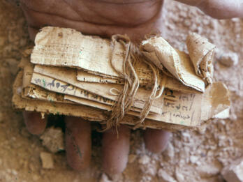 Close-up of a hand holding an ancient, rolled bundle of papyrus tied with string, resting on a dusty surface with small rocks or pieces of debris scattered around.