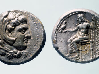 Front and back of an ancient silver coin. One side shows a profile of a man's head with curly hair, while the other side depicts a seated figure holding a bird, with Greek inscriptions beside the figure.
