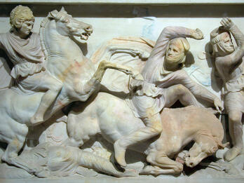 A marble relief sculpture shows three figures in battle: a mounted warrior attacks another figure who is falling from a horse, while a third figure takes a defensive pose. Two additional figures, apparently dead, lie on the ground.