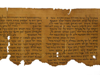 A fragment of an ancient, weathered scroll with rows of Hebrew text. The parchment edges are torn and uneven, showing signs of age and wear.
