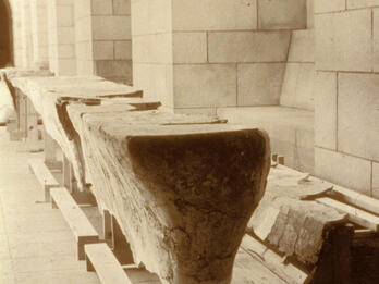 Wooden supports in the shape of a long, narrow table and bench with slabs of broken plaster on top, in a room with arched ceilings and stone walls.