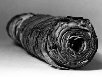 A black and white photo of an ancient, rolled-up scroll with layers of worn, textured material visible, resting on a plain surface.