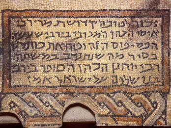Ancient mosaic with Hebrew script, bordered by geometric patterns, including interwoven designs at the bottom. The text is clearly visible within a rectangular frame.