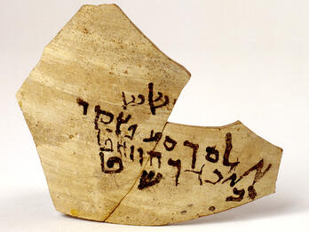 A broken piece of pottery with ancient Hebrew script written in black ink across its surface.