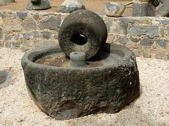A large, round olive press consisting of a circular basin and wheel sits outdoors on gravel, surrounded by a low stone wall and other stone objects.