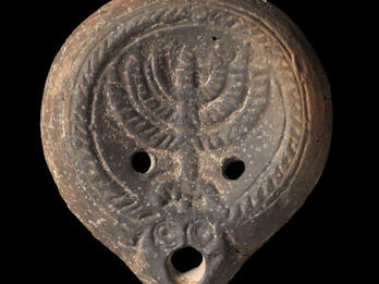 Ancient oil lamp with a decorative border and a central menorah design, with two holes flanking the menorah and a larger hole at the spout.