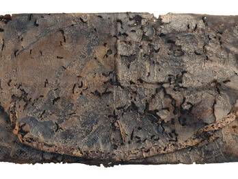 A folded rectangular piece of weathered, decayed leather with holes, dark stains, and a frayed strap or cord along one edge.