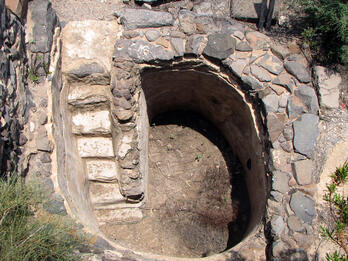 A round, stone-lined pit with built-in stone steps leading down to a dirt floor. The structure is surrounded by rocks and sparse vegetation.