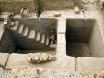 A person stands in a large, rectangular, stone excavation site with two deep pits, one with carved steps descending into it. The surrounding area is dry and sandy, with some scattered rocks.