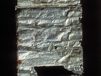 A fragment of an ancient, partially damaged silver scroll with faint inscriptions. Parts of the scroll are torn and missing.