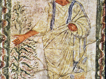 Ancient fresco shows a bearded man in a white robe with blue stripes, standing beside a tree, extending one arm outward. The background features simple landscape shapes, and a wavy border surrounds the scene.