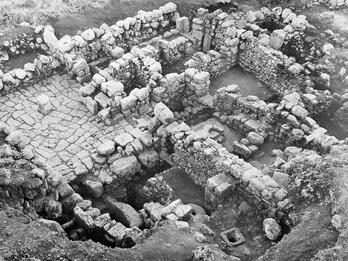 Aerial view of stone ruins, showing multiple rectangular rooms and passageways with uneven stone walls and floors, partially surrounded by earth and vegetation.