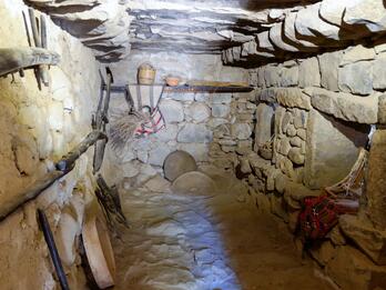 A small, narrow stone room with wooden shelves, woven baskets, pottery, and other household items. One wall has low window openings.