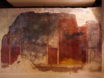 Ancient wall fresco with faded red, brown, and dark blue panels, partially damaged and surrounded by rough, exposed stone edges. The mural features simple geometric shapes and vertical divisions.