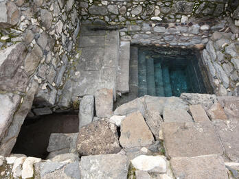 A stone structure with steps leading down into a rectangular pool of water, surrounded by weathered stone walls and blocks.