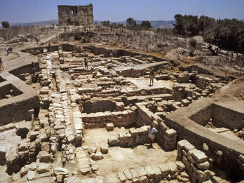 Ancient stone ruins with several people exploring the area; a partially intact building stands in the background under a clear blue sky.